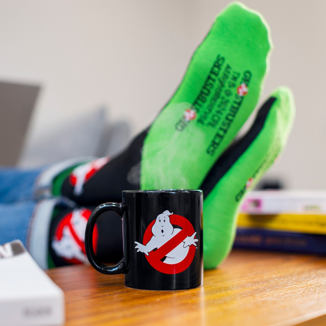 Czarny kubek z logo Ghostbusters stoi na drewnianym stole, za nim zielone skarpetki Ghostbusters.