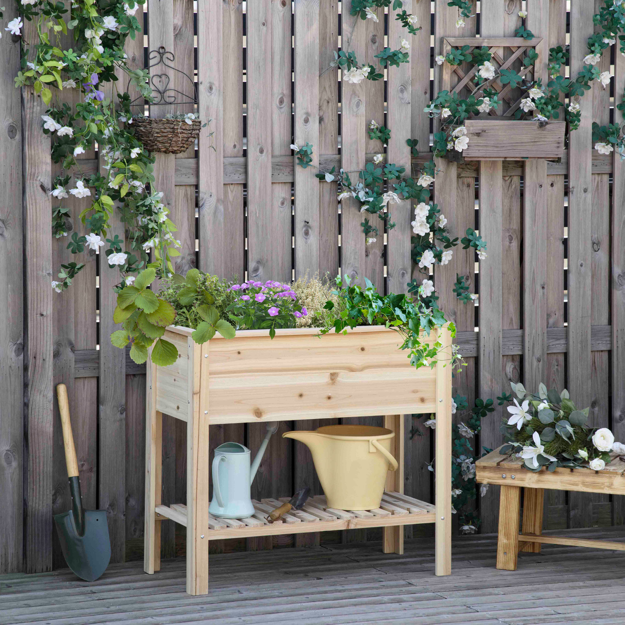 Un bancal elevado de madera con herramientas y flores contra una valla de madera.