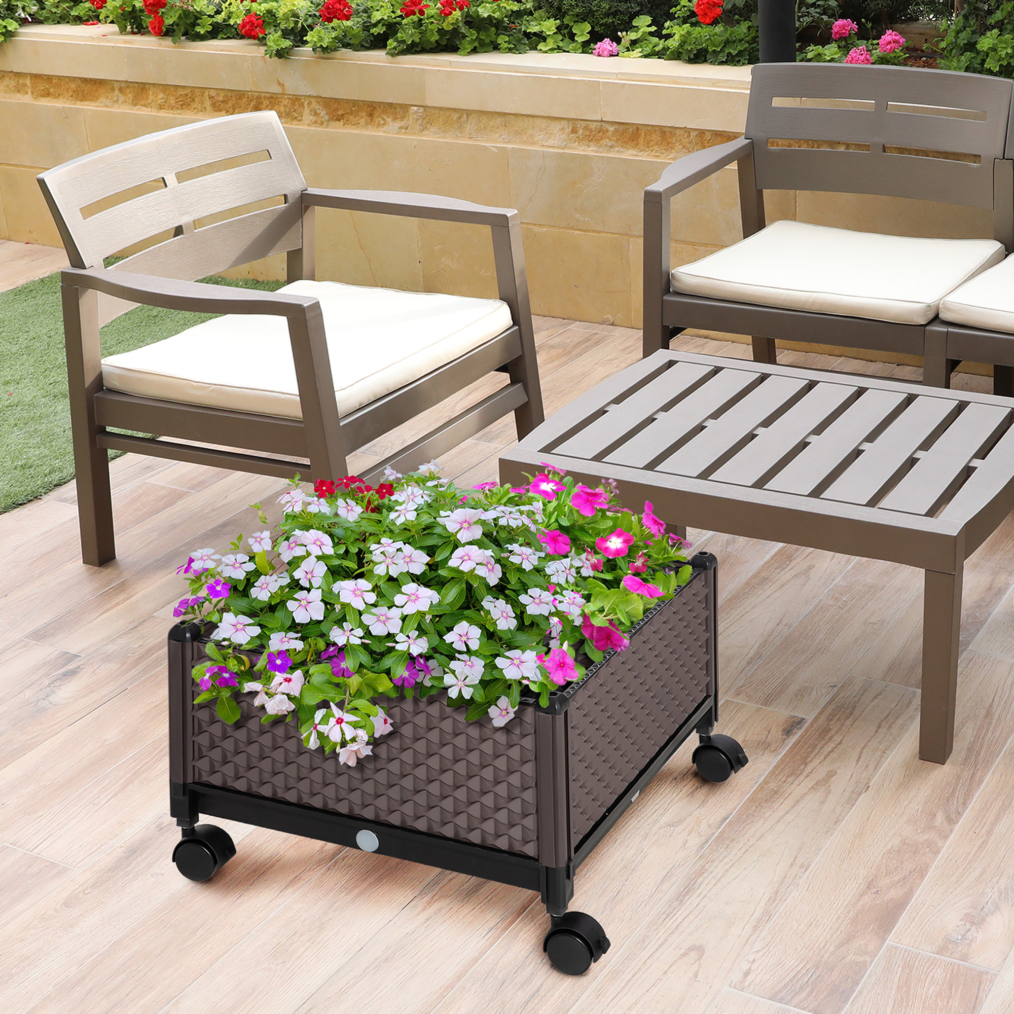 Una jardinera marrón con flores coloridas sobre ruedas está en una terraza de madera junto a muebles de jardín.