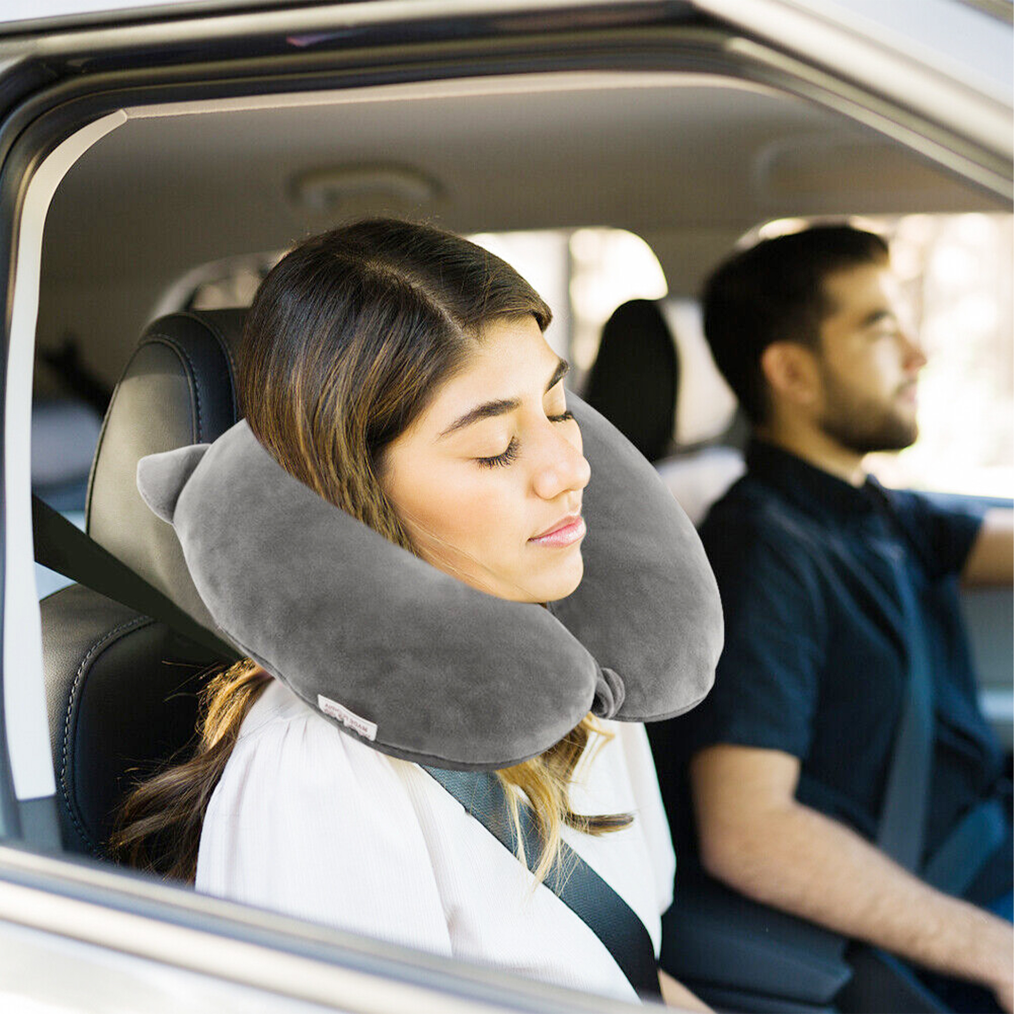 Donna dorme in auto con cuscino da viaggio, uomo guida sullo sfondo.