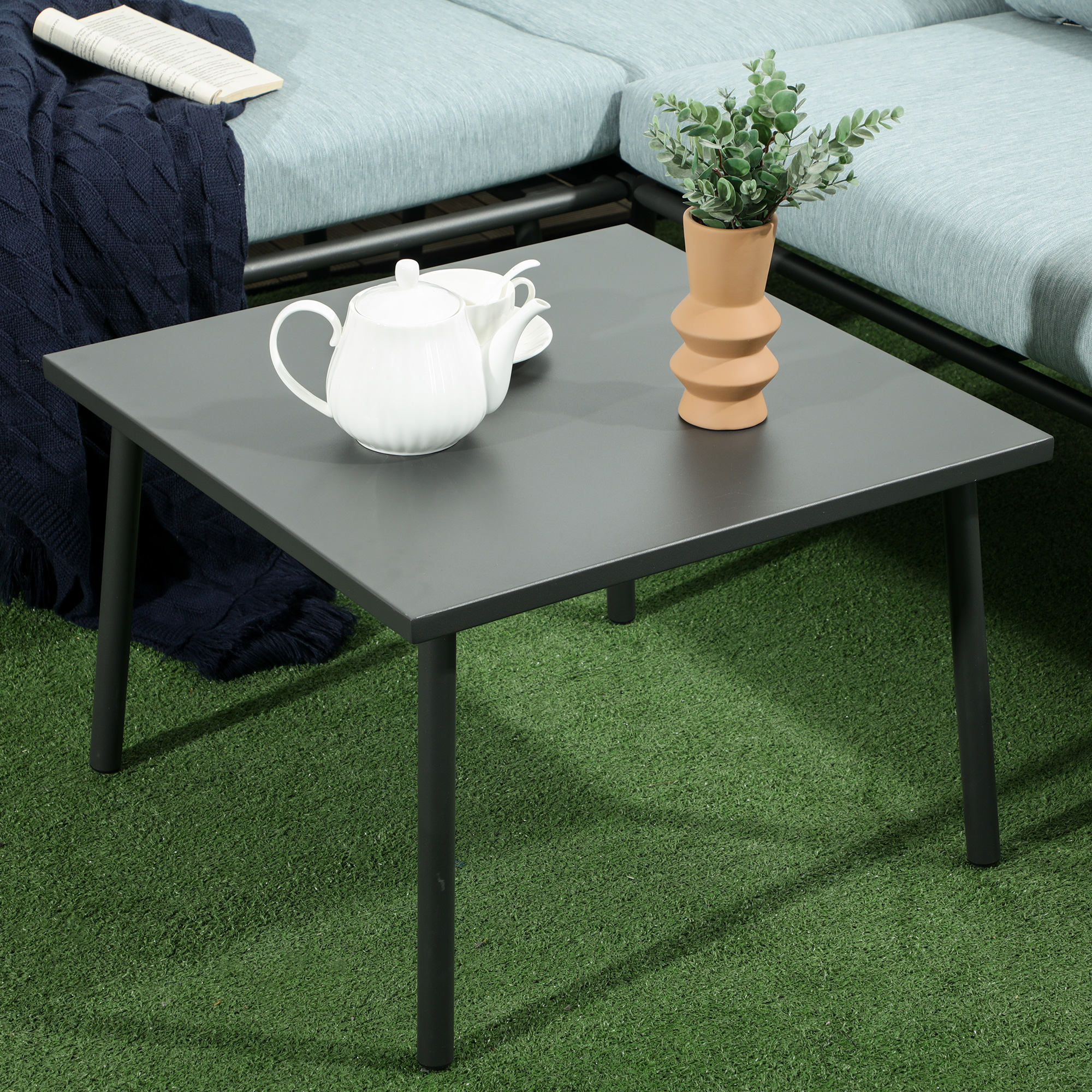 Un tavolo grigio scuro su erba verde con teiera bianca, tazza, pianta e coperta blu.