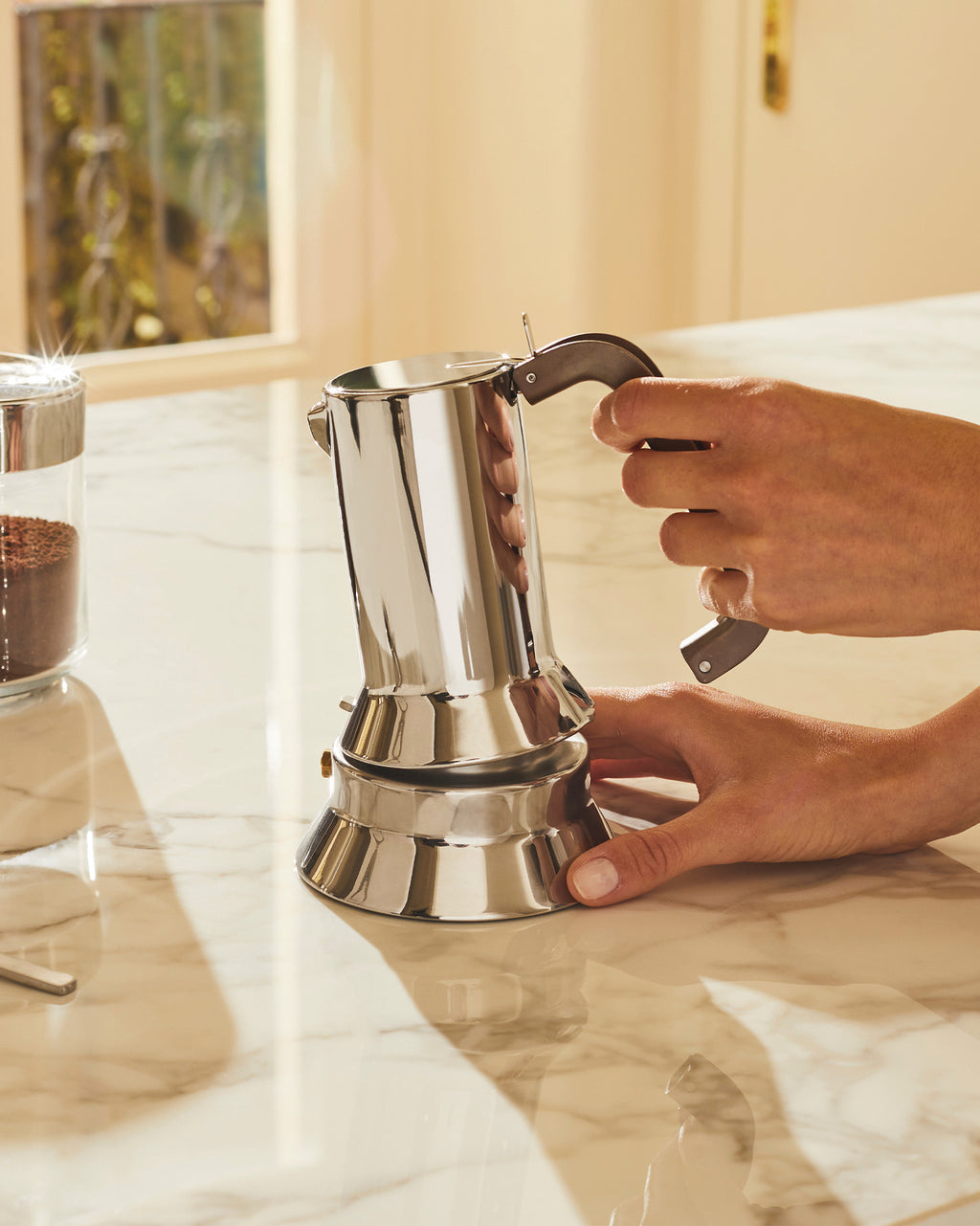 Person hält glänzende Mokkakanne auf Marmortisch. Kaffeepulver im Glas.