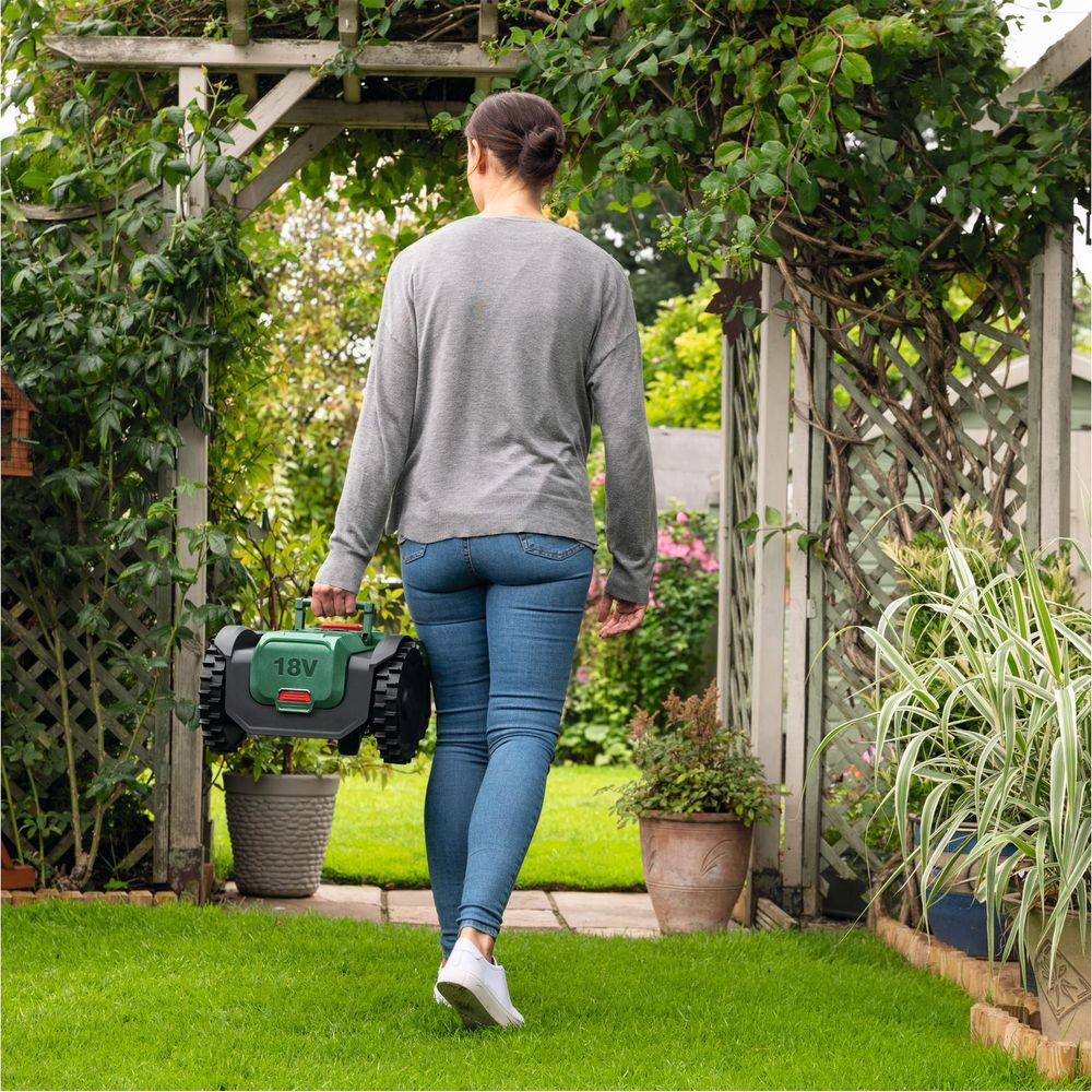 Una donna porta un piccolo robot tagliaerba verde in un giardino.