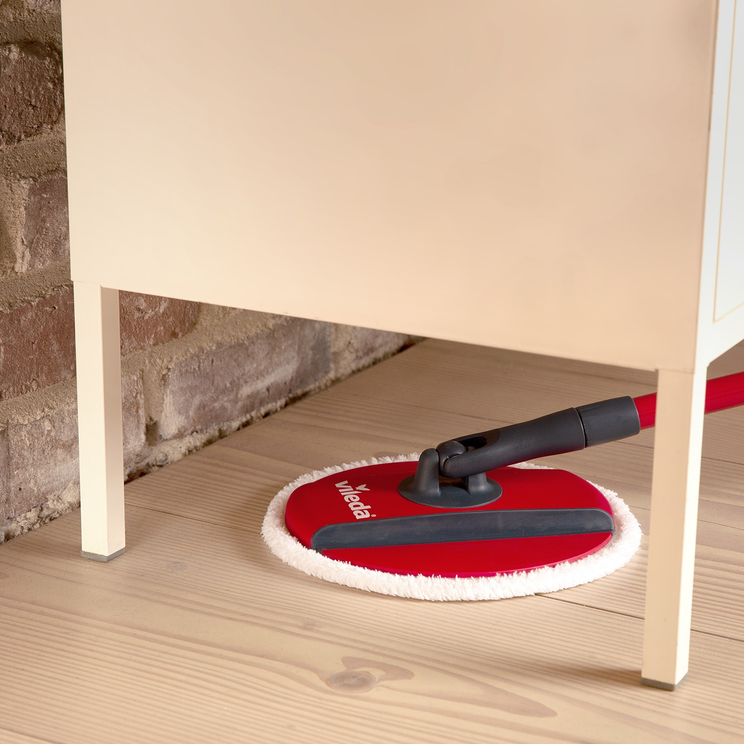 Un mocio Vileda rosso pulisce il pavimento in legno sotto un mobile chiaro.