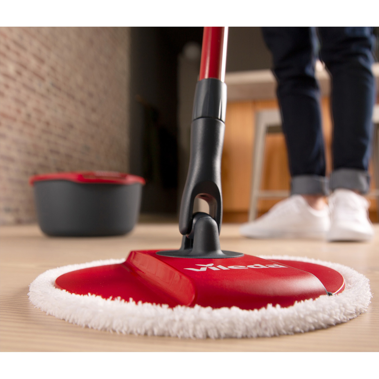 Una testa di mop rossa con frange bianche su pavimento in legno, un secchio vicino.