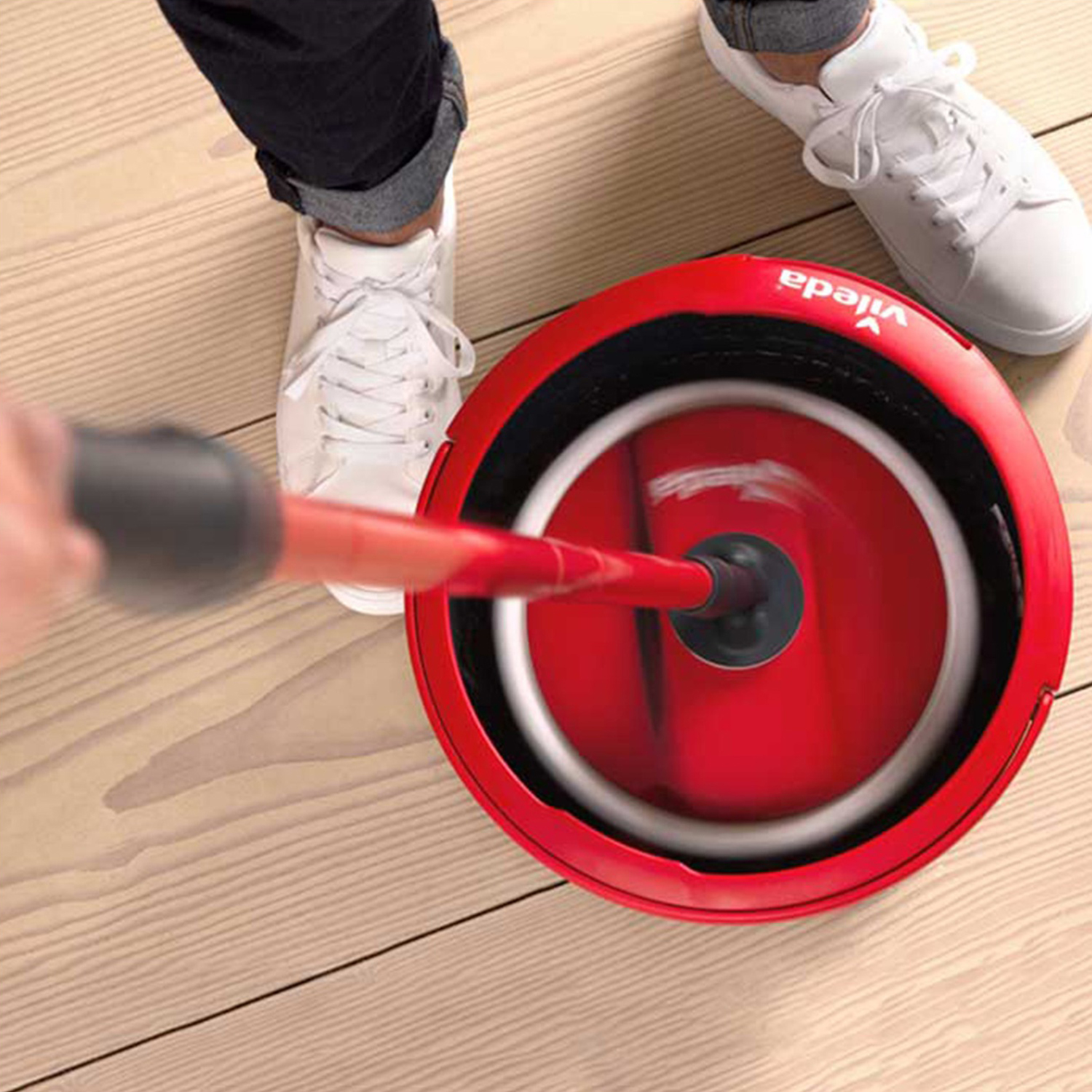 Un secchio per mocio rotante rosso con scarpa bianca e gambe su pavimento in legno.