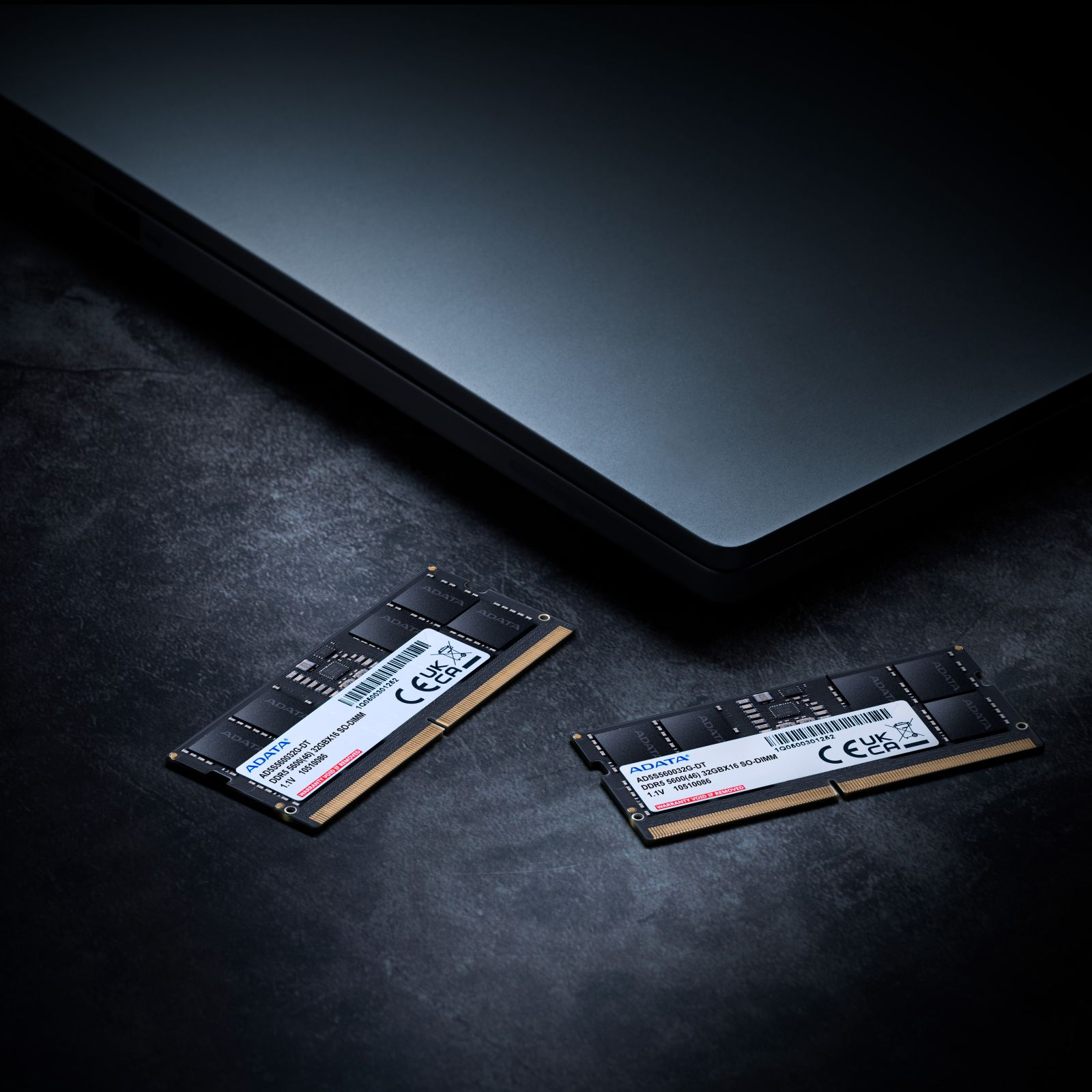Zwei schwarze RAM-Module liegen auf dunklem Grund neben einem Laptop.