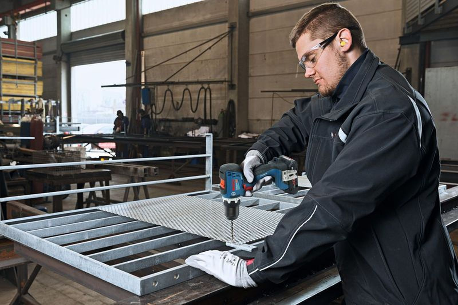 Mann bohrt in Werkstatt in Metallgitter, trägt Schutzbrille und Handschuhe.