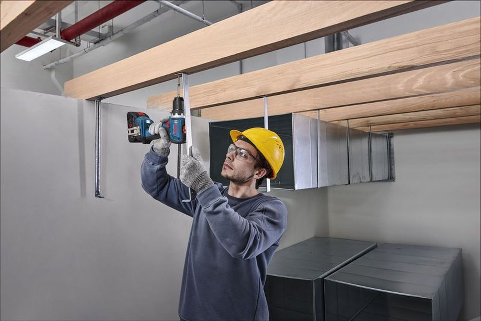 Ein Mann mit Helm bohrt Holzträger. Lüftungskanäle sind sichtbar.