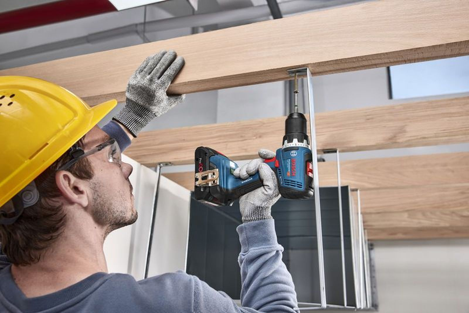 Mann mit Bohrer schraubt Holz in die Decke. Trägt gelben Helm und Handschuhe.