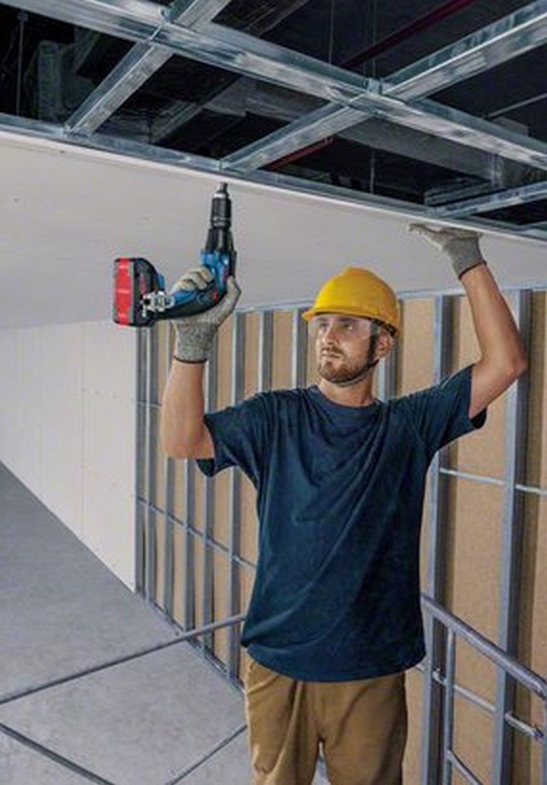 Un uomo con elmetto giallo fora una piastrella del soffitto in una struttura metallica.