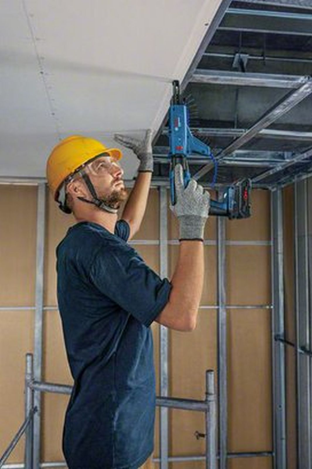 Un operaio edile con casco giallo installa cartongesso al soffitto con avvitatore.