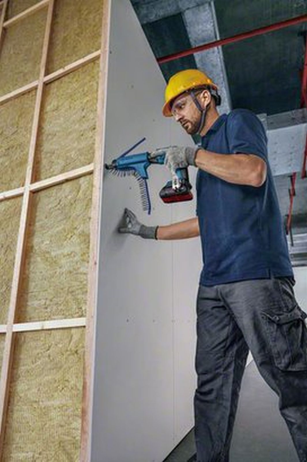 Uomo con elmetto avvita cartongesso a montanti in legno con avvitatore.