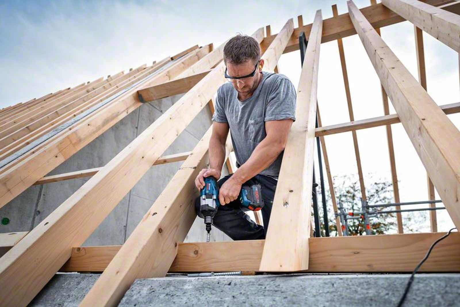 Mann im grauen Hemd und Schutzbrille bohrt mit blauer Bohrmaschine Holzdachbalken.