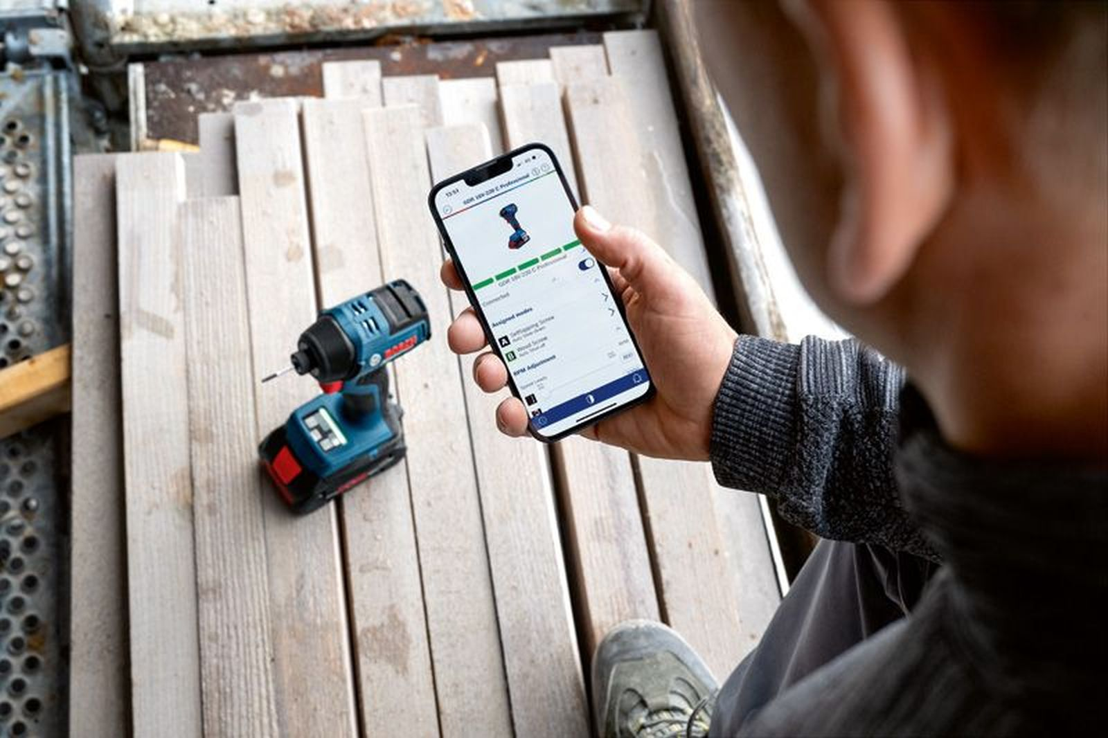 Ein Mann hält ein Smartphone mit einer Bohrmaschinen-App. Daneben liegt eine blaue Bohrmaschine.