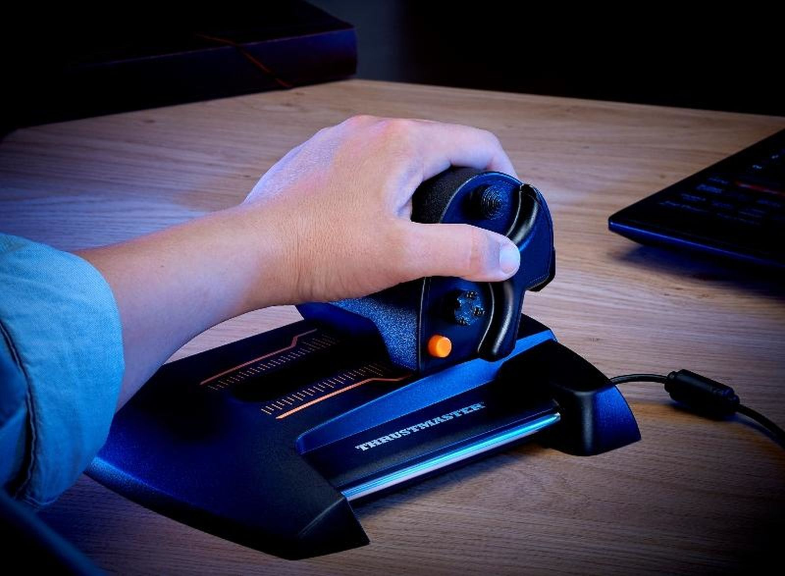 Une main tient un joystick noir avec des accents bleus sur un bureau en bois.