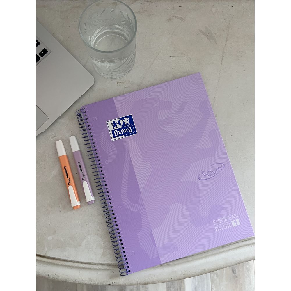 Ein lila Oxford Notizbuch mit zwei Stiften, einem Glas Wasser und einem Laptop.