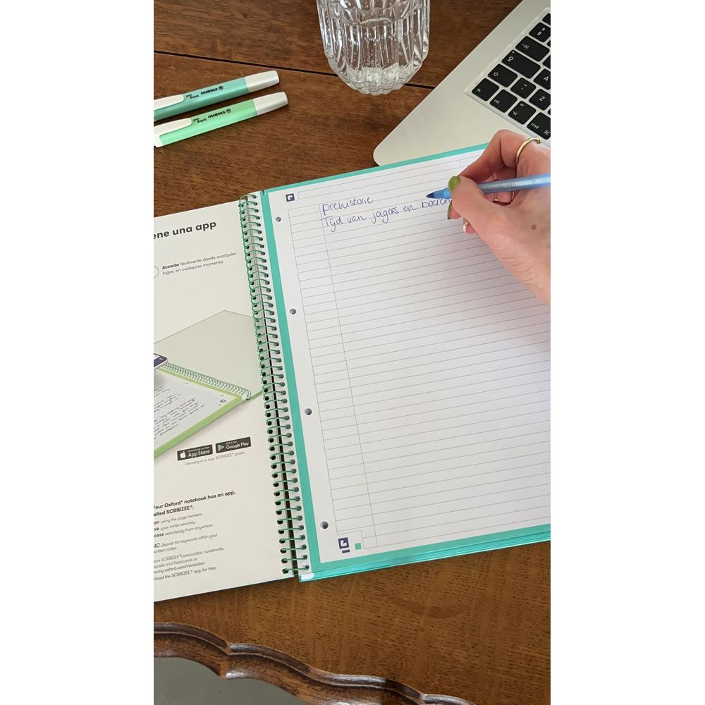 Eine Hand schreibt in ein Notizbuch. Stifte, Laptop und Glas auf dem Tisch.