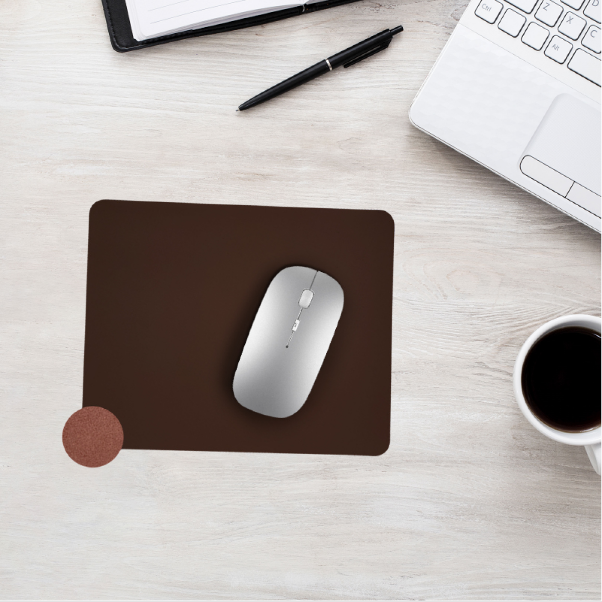 Silberne Maus auf brauner Unterlage, neben Kaffee, Laptop, Stift und Notizbuch.