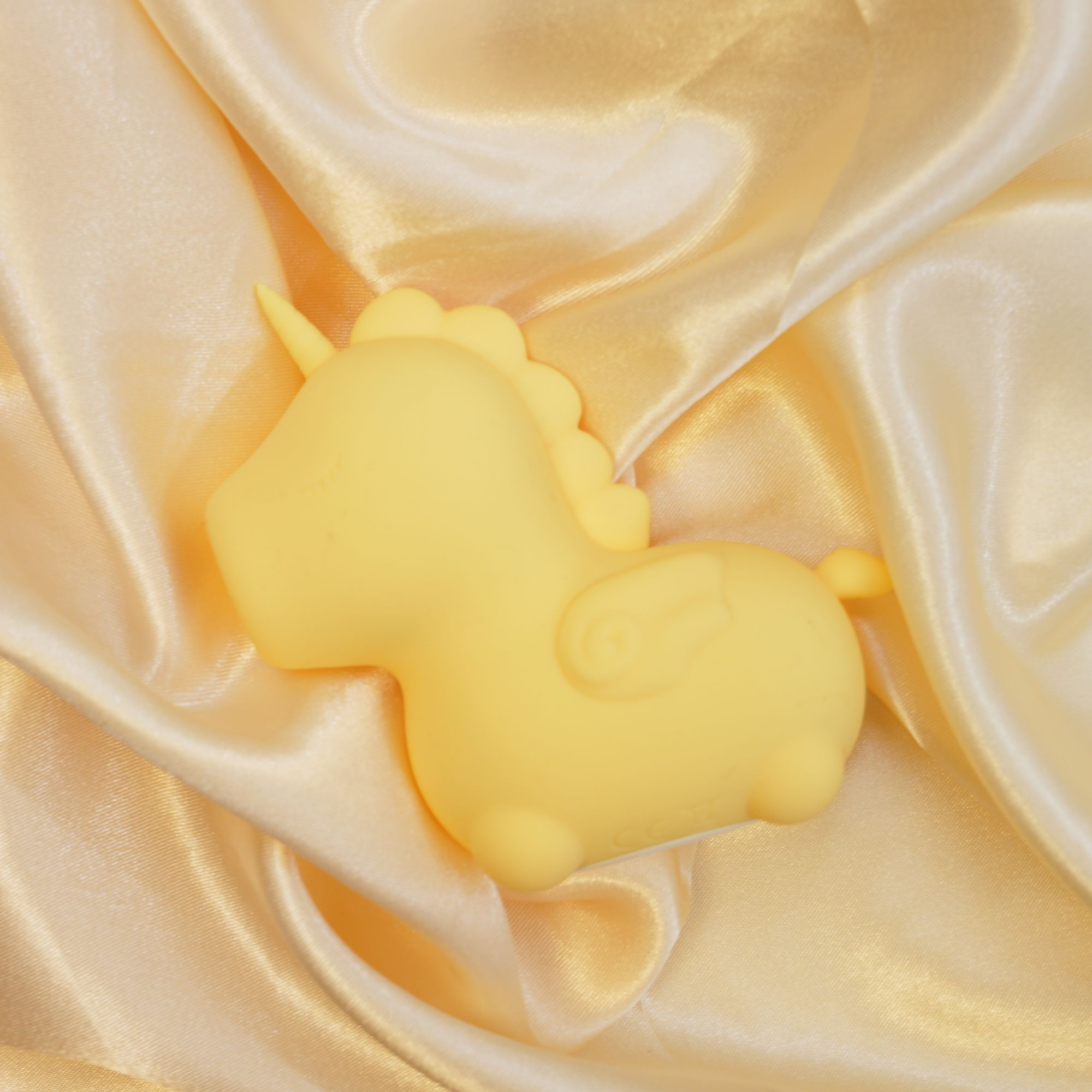 Ein gelbes Einhorn-Spielzeug liegt auf glänzendem, cremefarbenem Stoff.