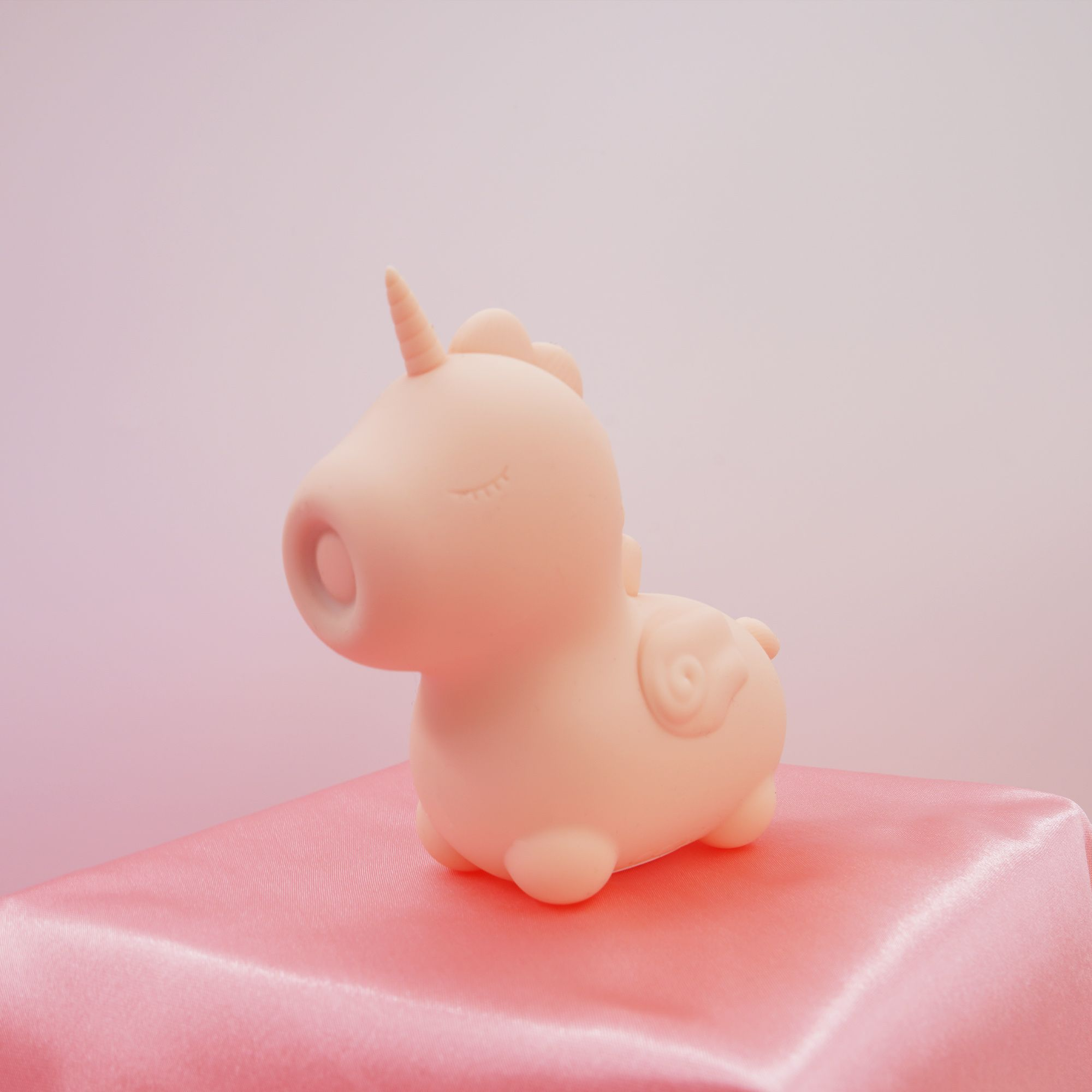 Ein pfirsichfarbenes Einhorn-Spielzeug mit geschlossenen Augen sitzt auf einem rosa Satintuch.