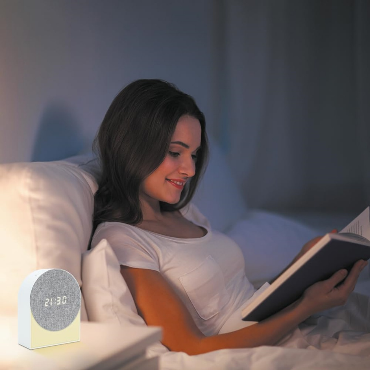 Eine Frau liest ein Buch im Bett mit einem digitalen Wecker im Vordergrund.
