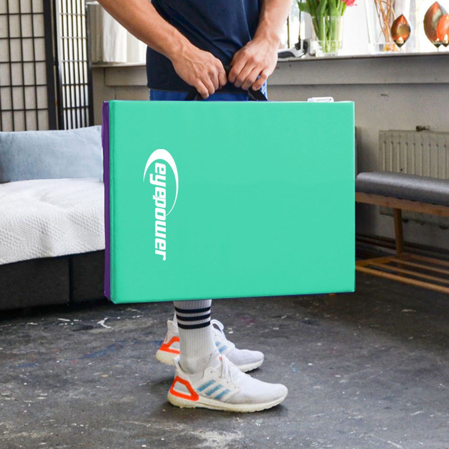 Ein Mann hält eine große, grüne Matte mit einem weißen Logo. Er trägt weiße Turnschuhe.