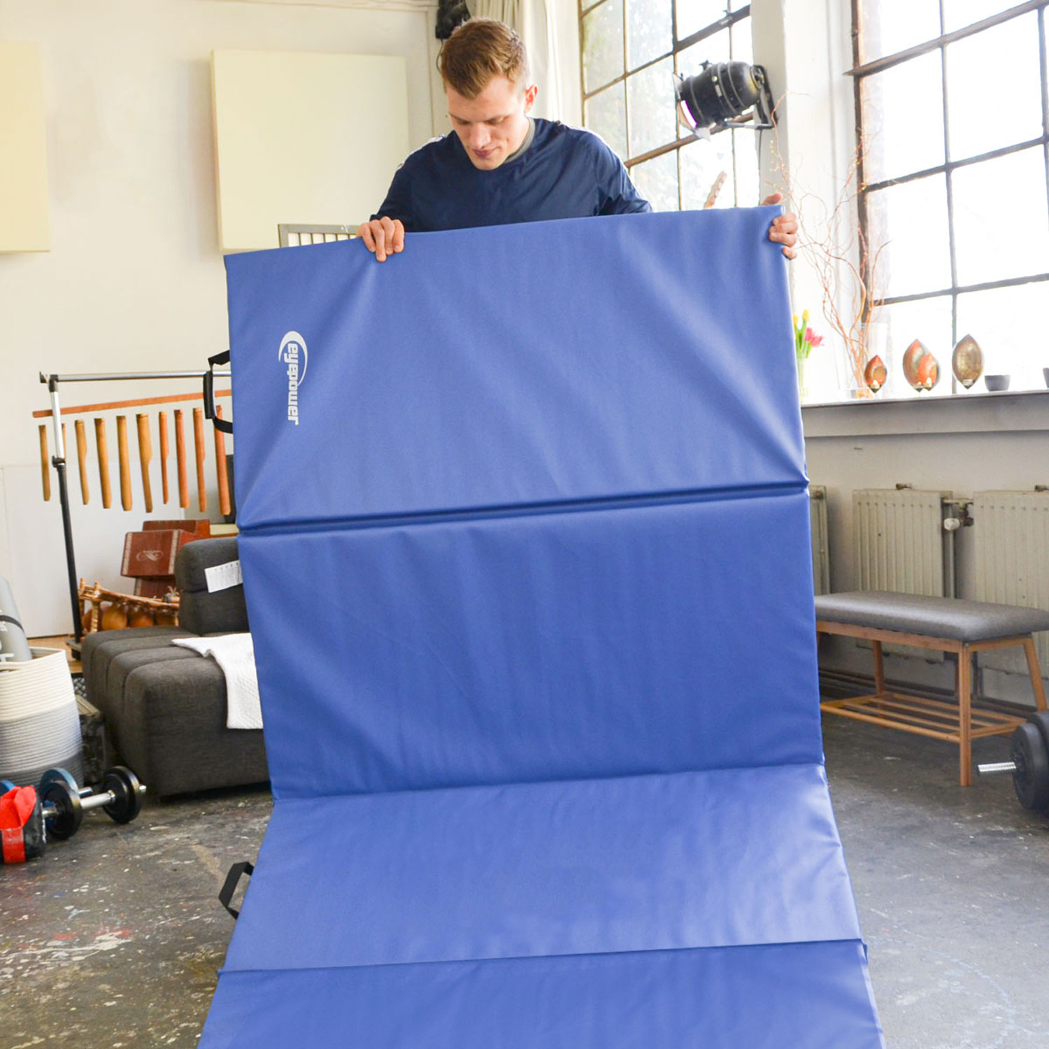 Ein Mann entfaltet eine blaue Matte in einem Raum mit Fenstern, Hanteln und einem Sofa.