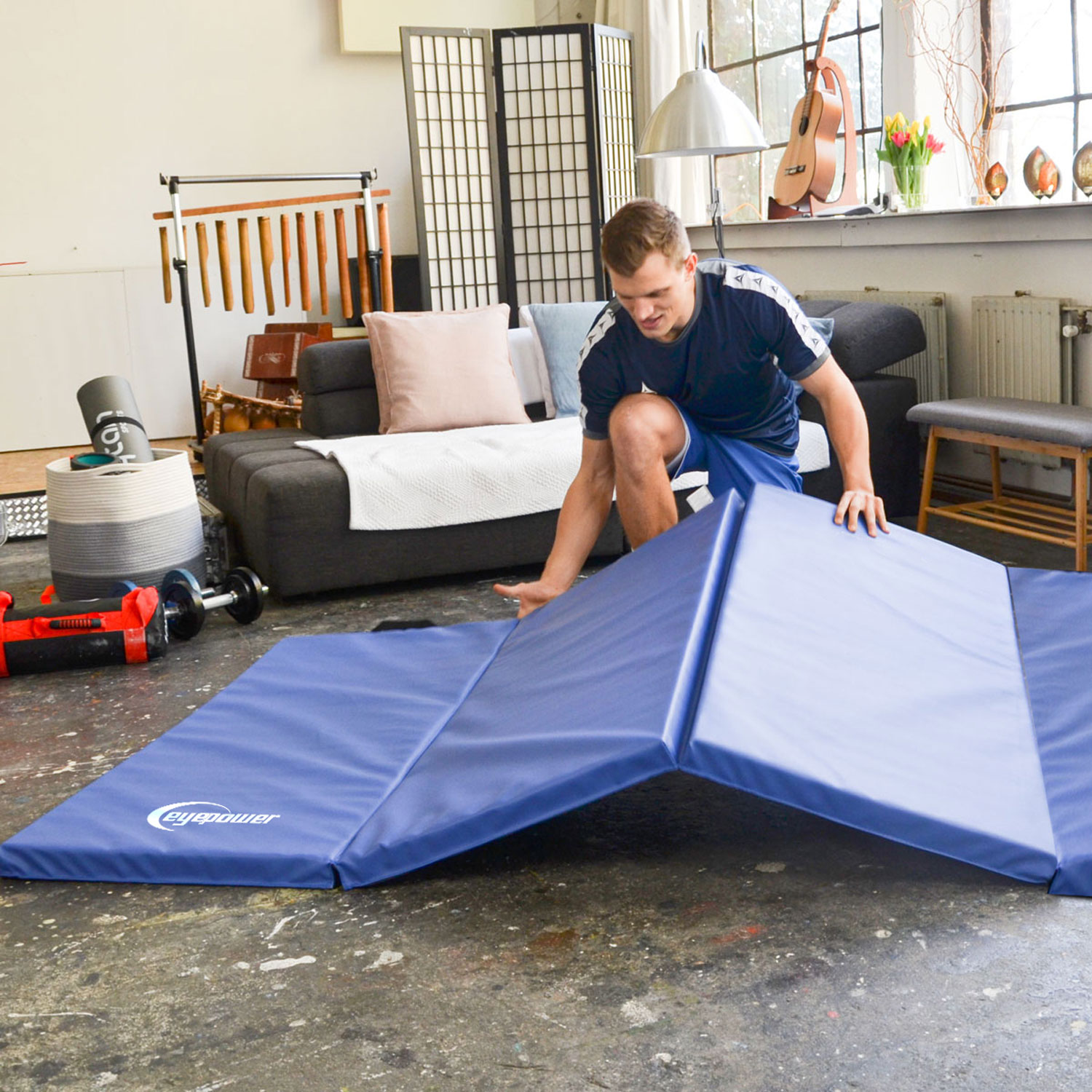 Ein Mann faltet eine blaue Gymnastikmatte in einem Wohnzimmer mit Möbeln und Fenstern.