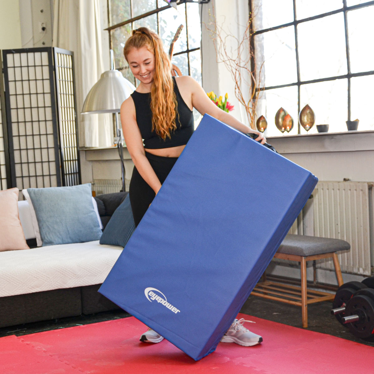 Eine Frau hält eine blaue Gymnastikmatte in einem Raum mit Fenstern und Möbeln.