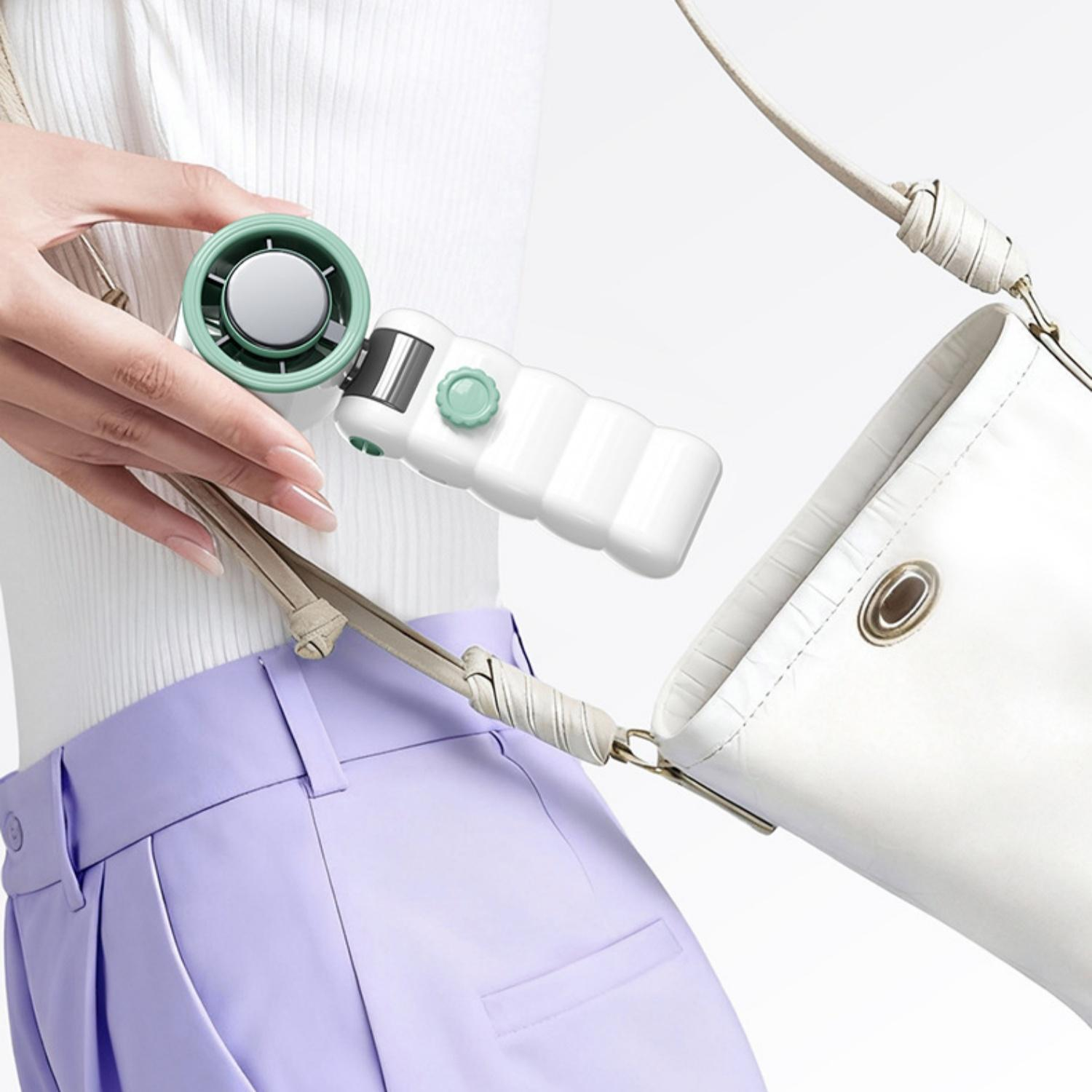 Eine Hand hält einen kleinen grünen Ventilator neben einer weißen Tasche und lila Hose.