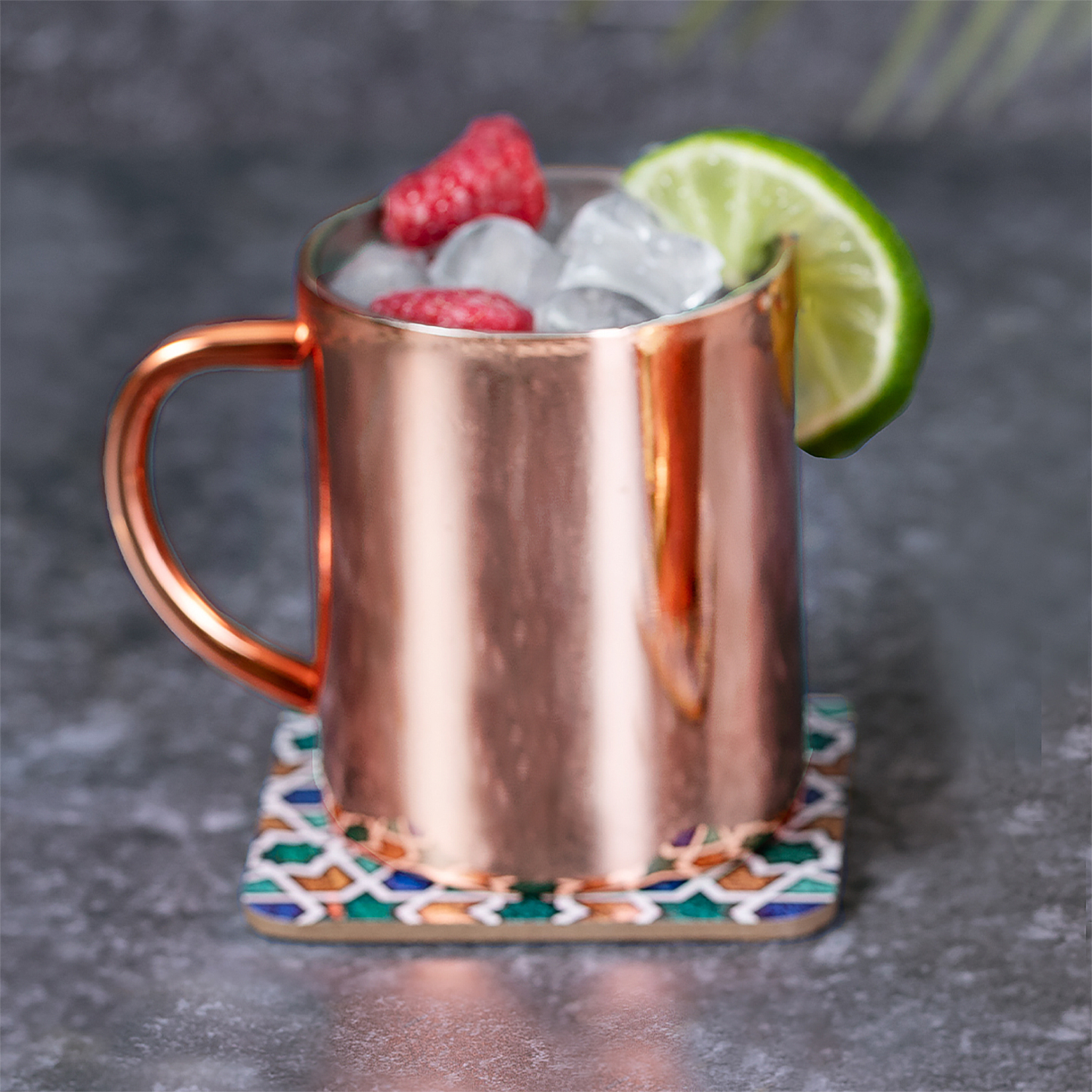 Una taza de cobre con hielo, bayas y lima sobre un posavasos colorido.