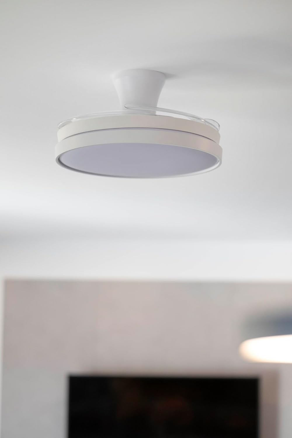 Un moderno ventilador de techo blanco con una lámpara circular está montado en un techo blanco, con una habitación borrosa debajo.