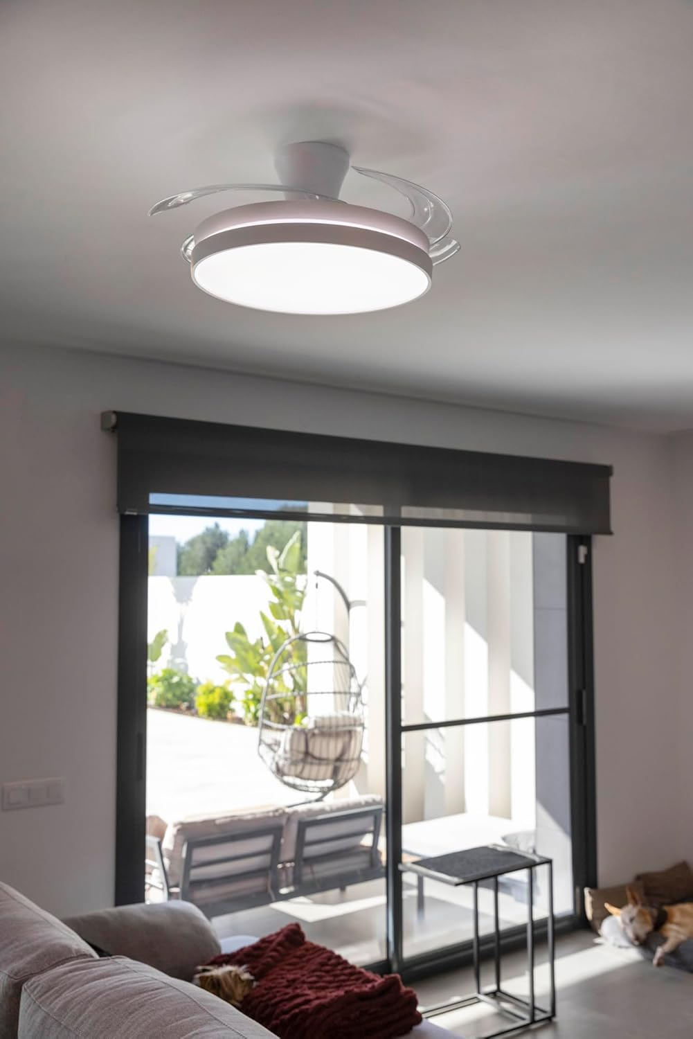 Un ventilador de techo con luz sobre un sofá con manta roja. Afuera se ve una silla colgante.