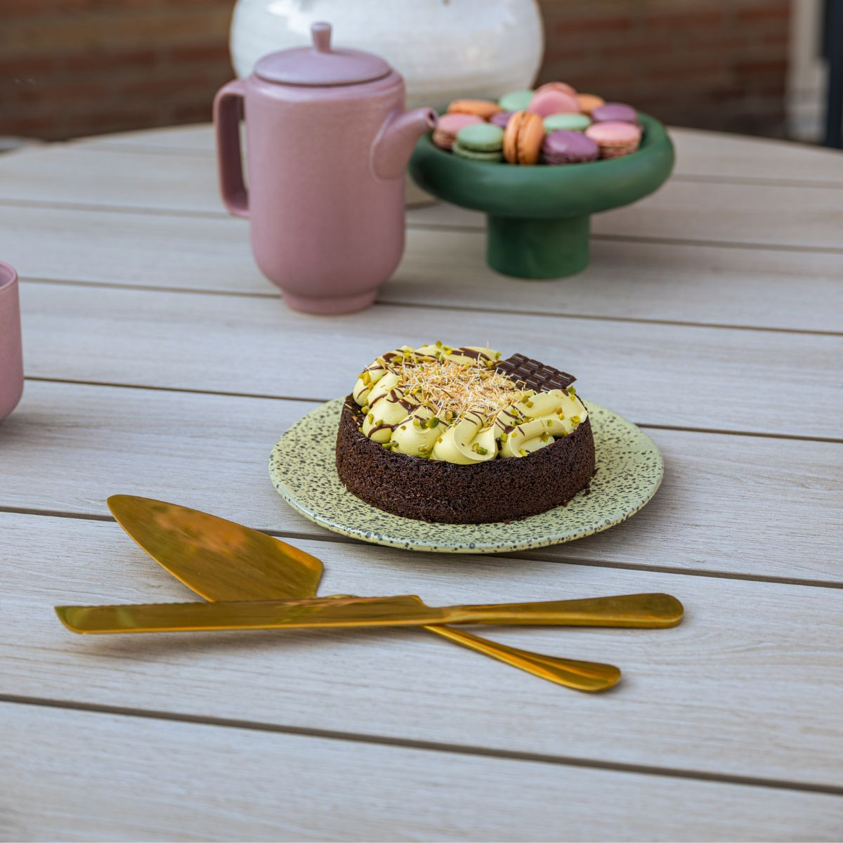 Chocoladetaart met gele room, pistachenoten en chocolade garnering, naast serveergerei.