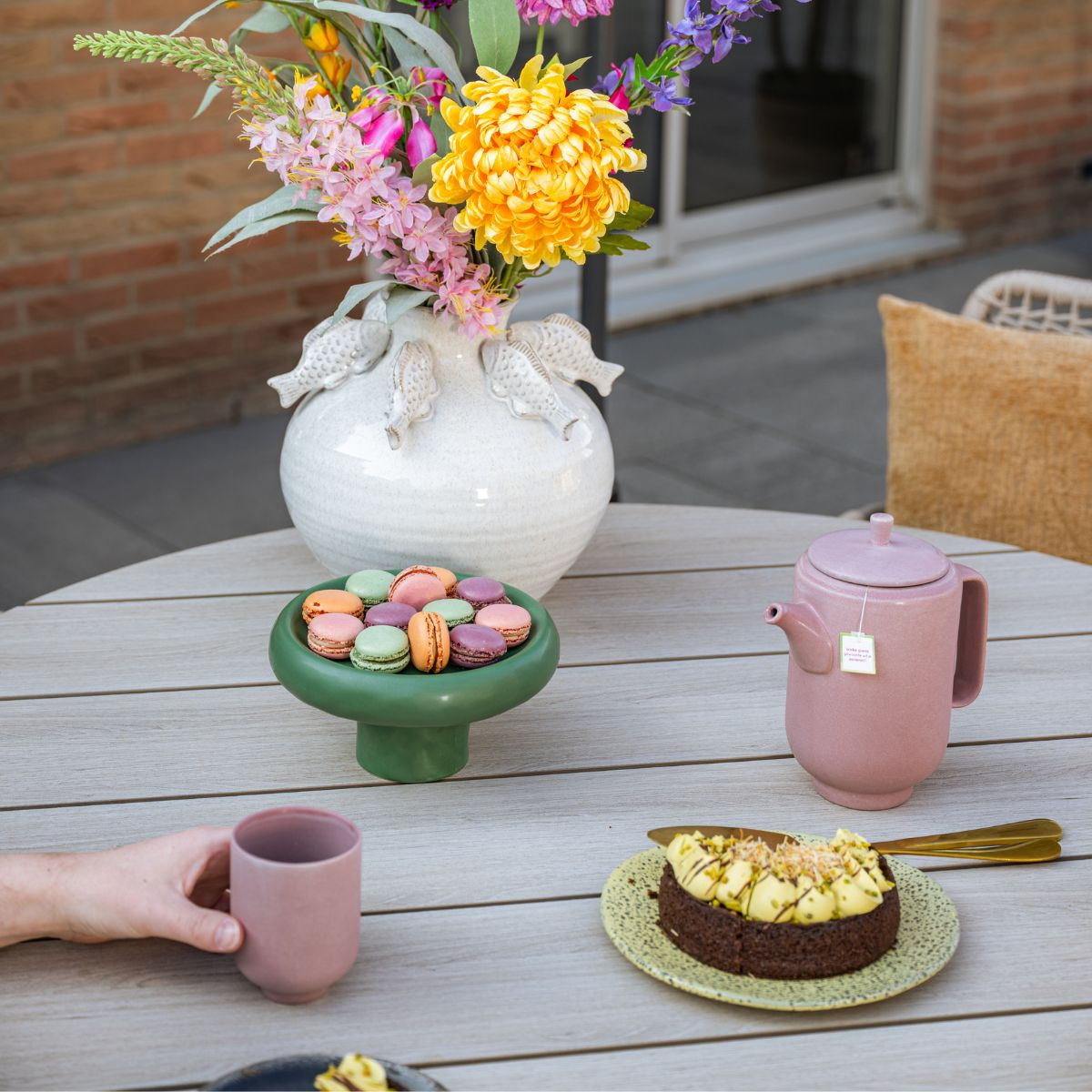 Een tafel met macarons, taart, theepot, vaas met bloemen en een hand die een kopje vasthoudt.