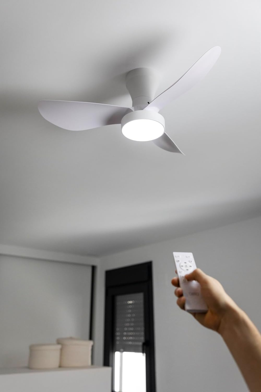 Una mano sostiene un mando a distancia blanco frente a un ventilador de techo blanco con luz.
