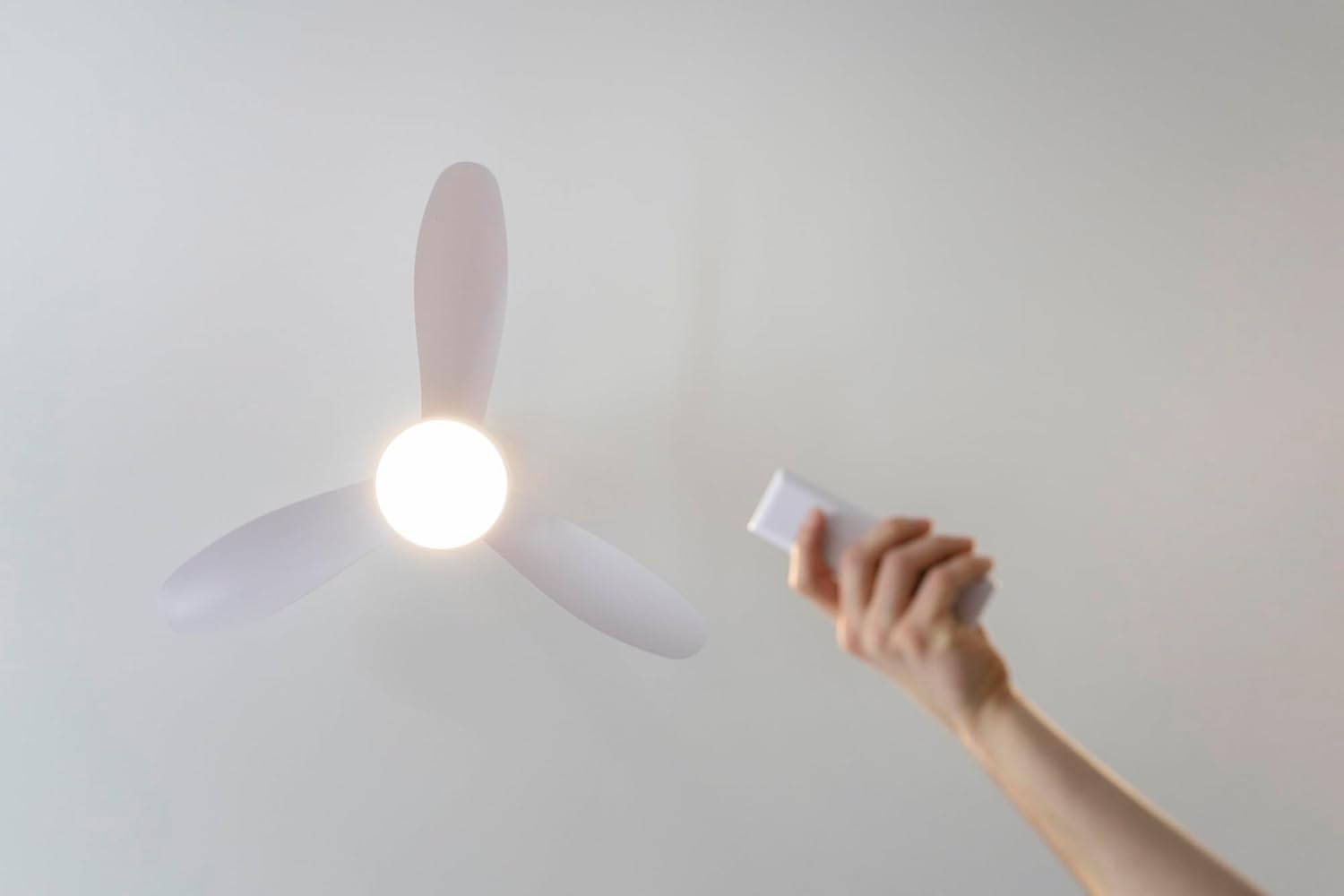 Una mano sostiene un mando a distancia junto a un ventilador de techo blanco con luz.