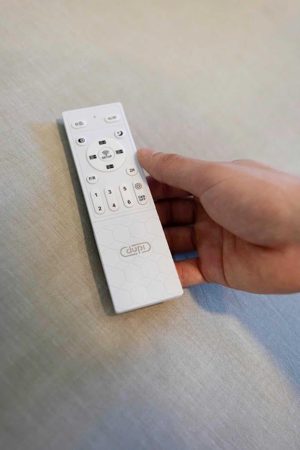 Una mano sostiene un mando a distancia blanco con botones numerados y un logo.