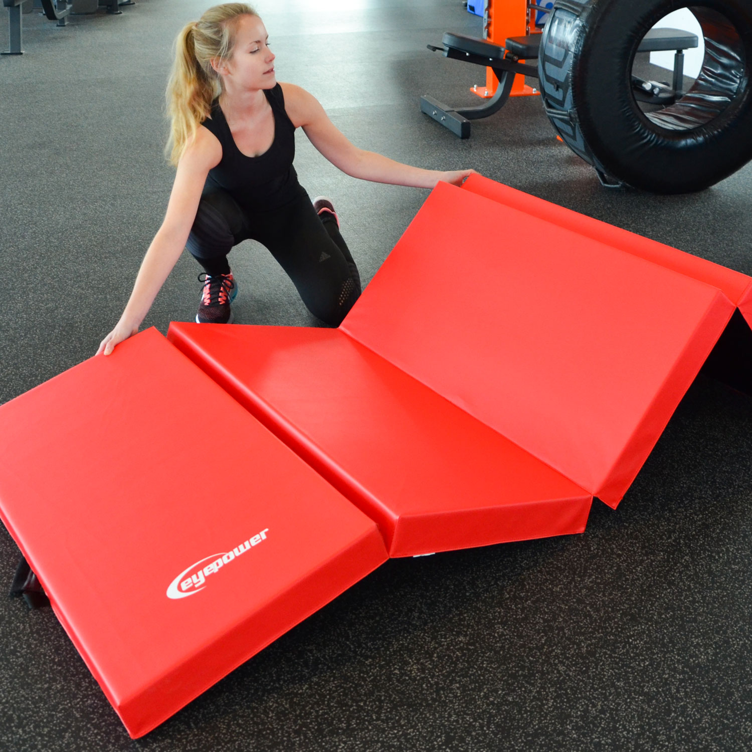 Frau in schwarzer Sportkleidung entfaltet rote Gymnastikmatten auf grauem Boden. Trainingsgeräte im Hintergrund.