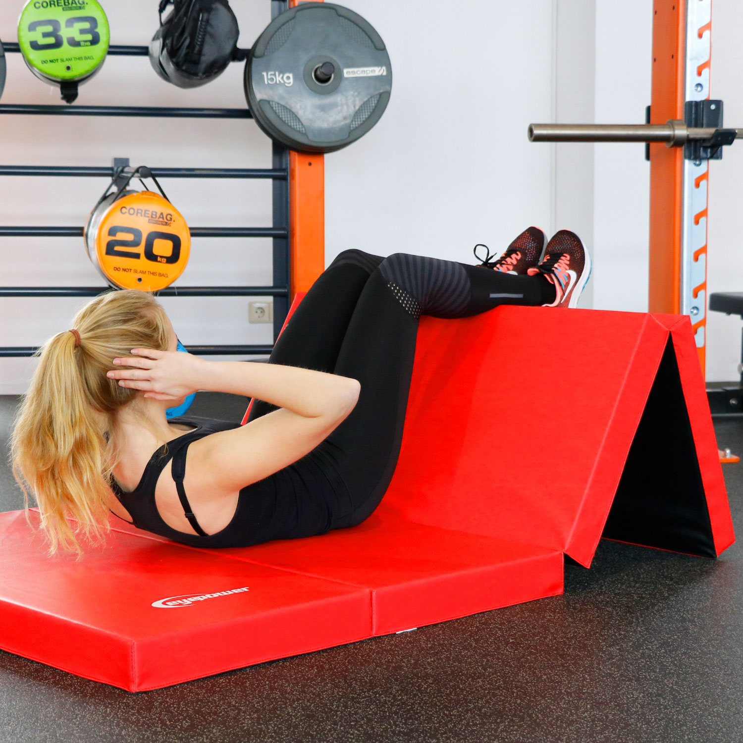 Frau macht Crunches auf einer roten Matte, im Hintergrund Hanteln und Corebags.