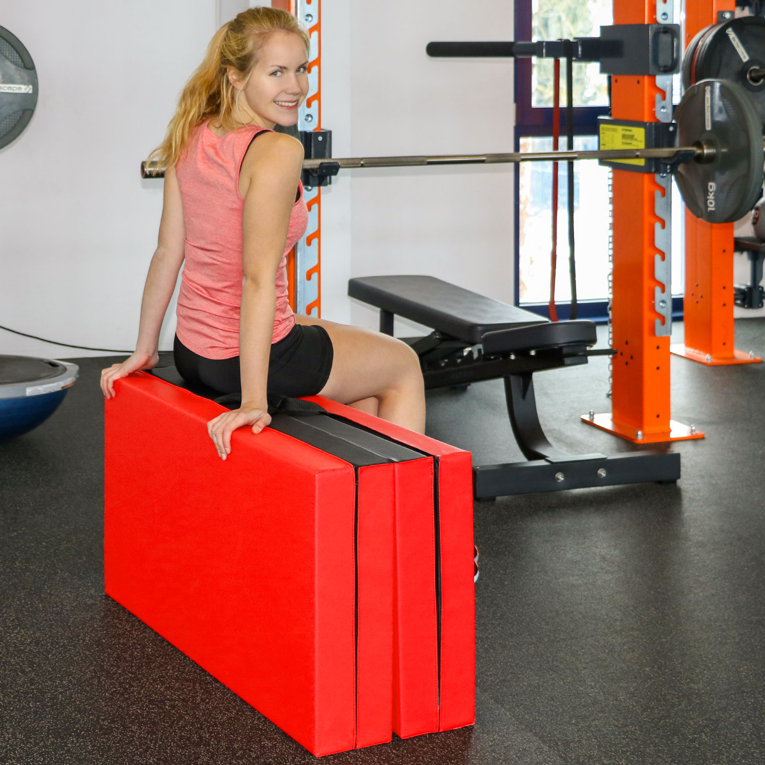 Eine Frau sitzt auf einer roten Matte in einem Fitnessstudio neben Hanteln und einer Hantelbank.