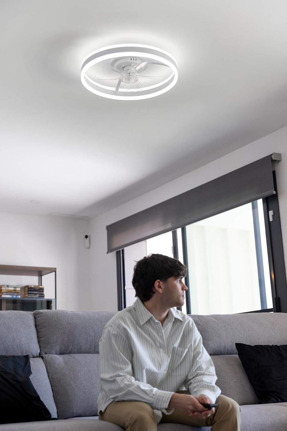 Un hombre sentado en un sofá bajo una lámpara de techo con ventilador, mirando por la ventana.