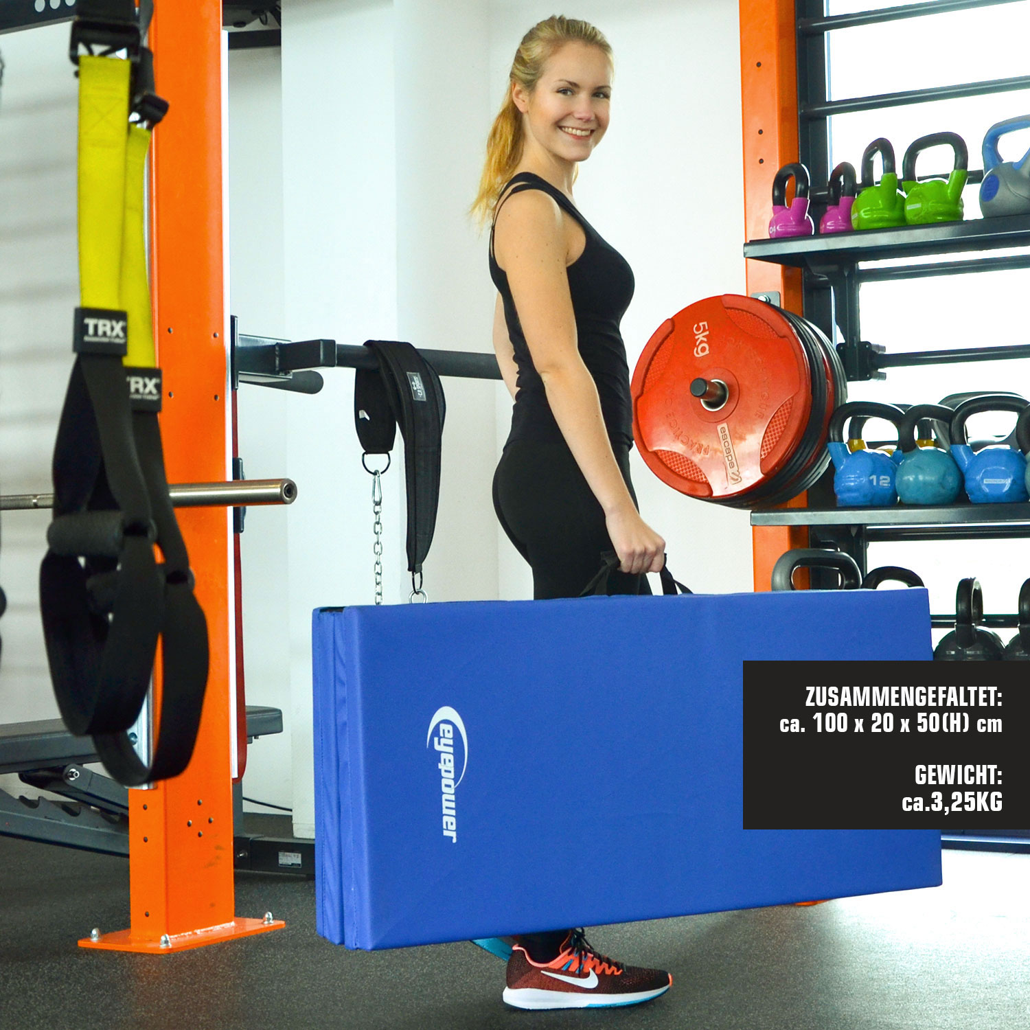 Eine Frau in Schwarz hält eine blaue Matte in einem Fitnessstudio mit Gewichten und TRX.