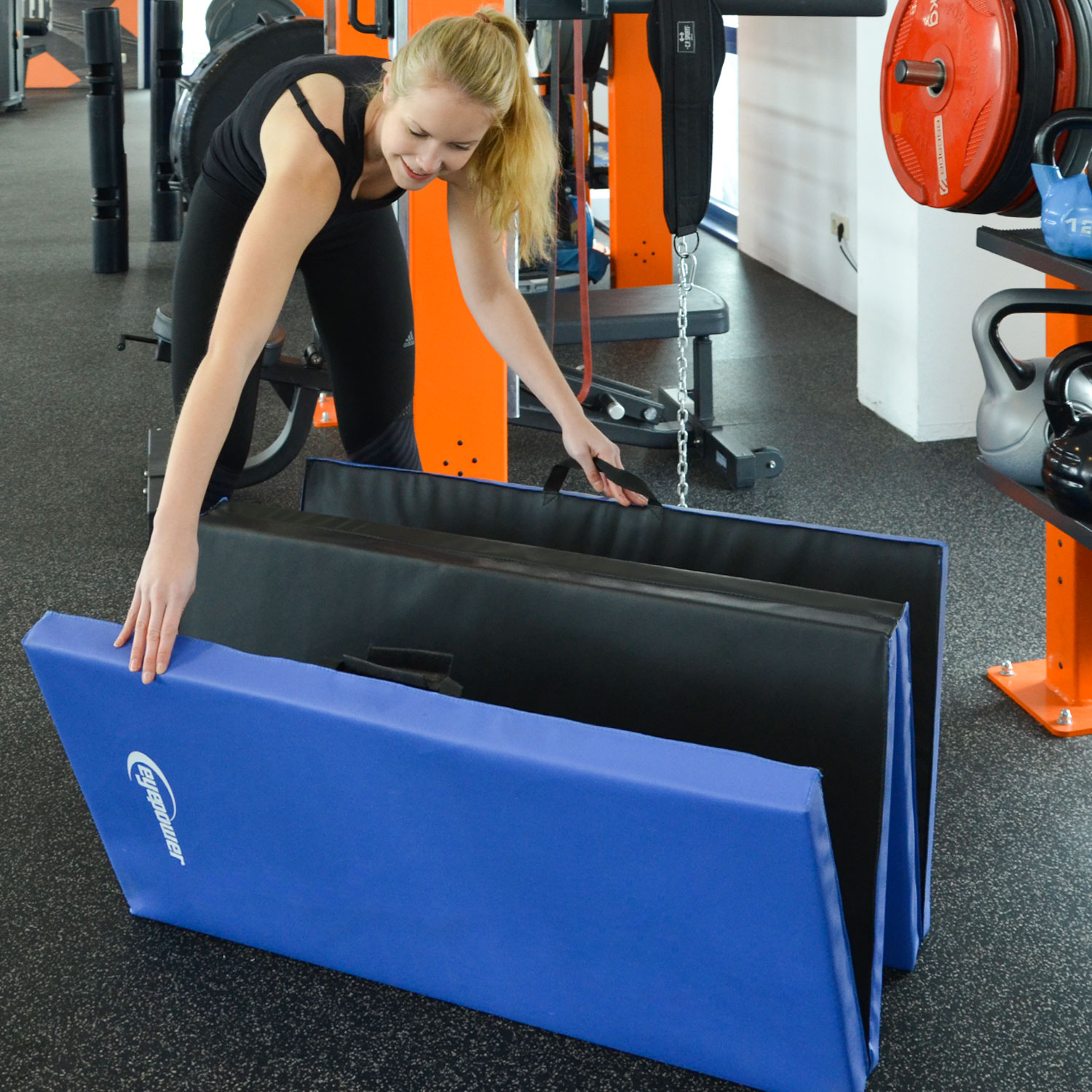Eine Frau faltet blaue und schwarze Matten in einem Fitnessstudio zusammen.