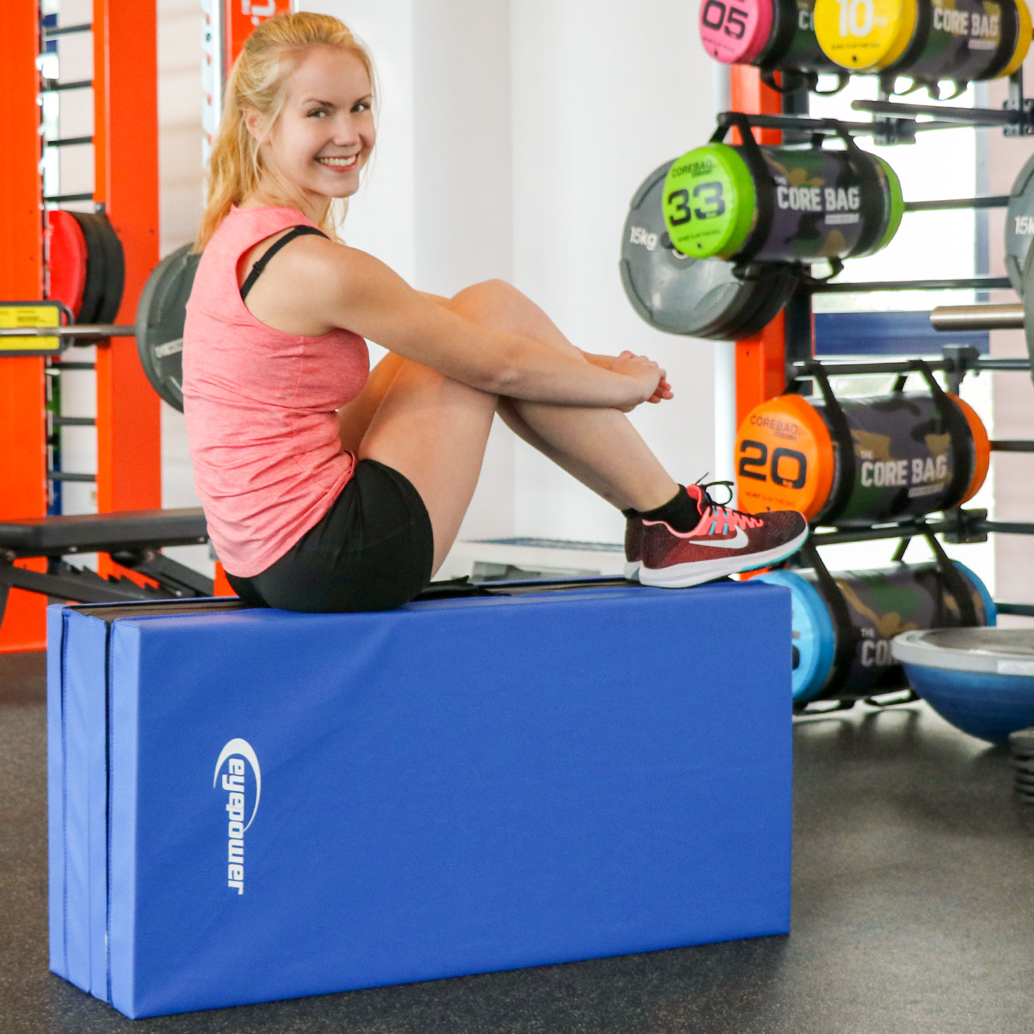 Eine Frau sitzt auf einer blauen Matte, Trainingsgeräte im Hintergrund.