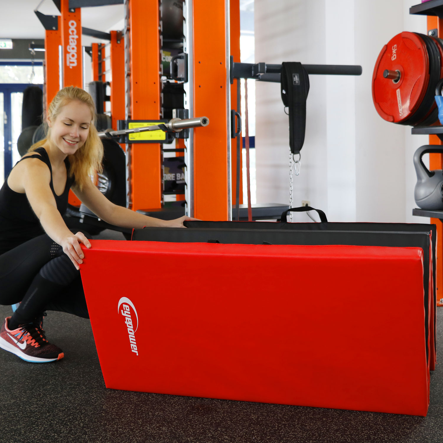 Eine Frau in Schwarz kniet vor roten und schwarzen Gymnastikmatten in einem Fitnessstudio.