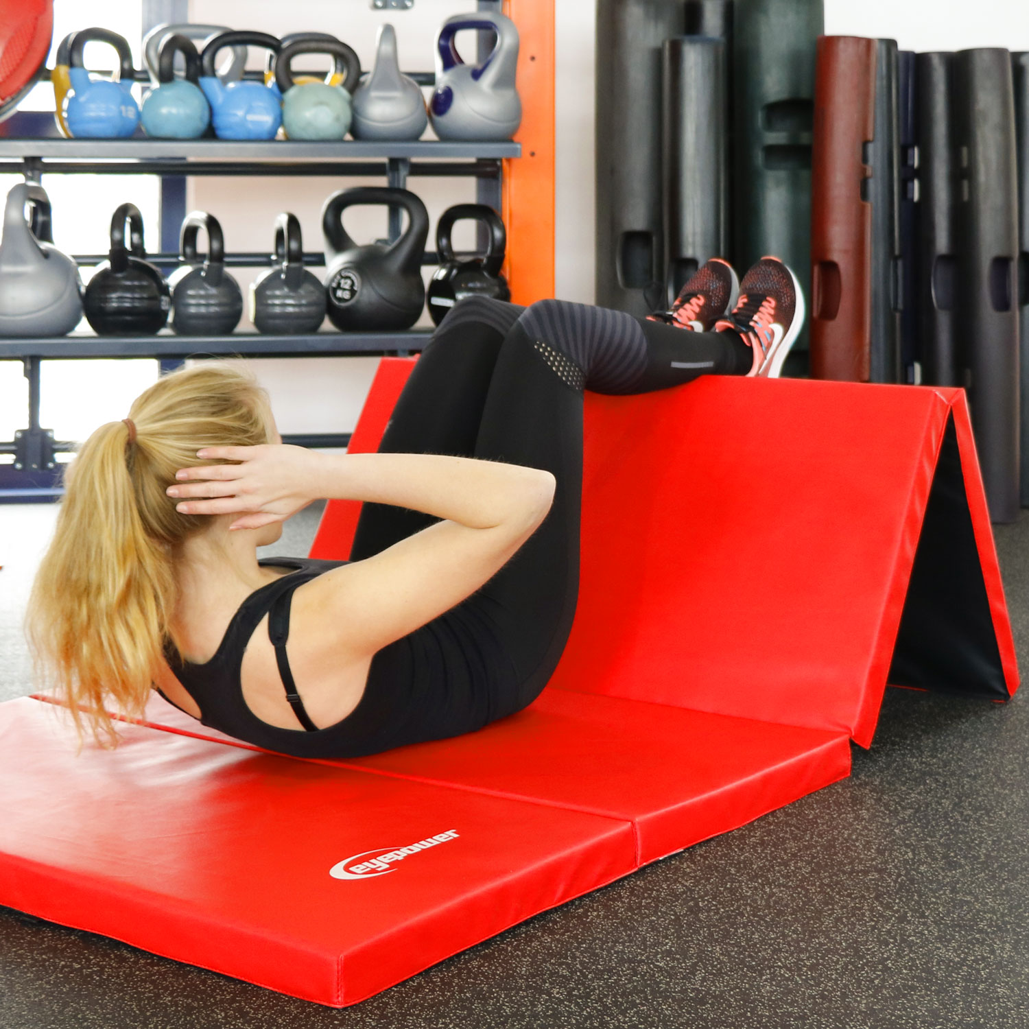 Frau macht Crunches auf einer roten Matte in einem Fitnessstudio mit Gewichten im Hintergrund.