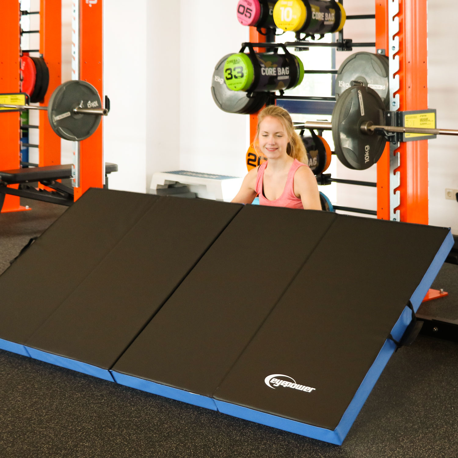 Eine Frau im Fitnessstudio lächelt vor einer ausklappbaren Matte, Gewichten und Core Bags.