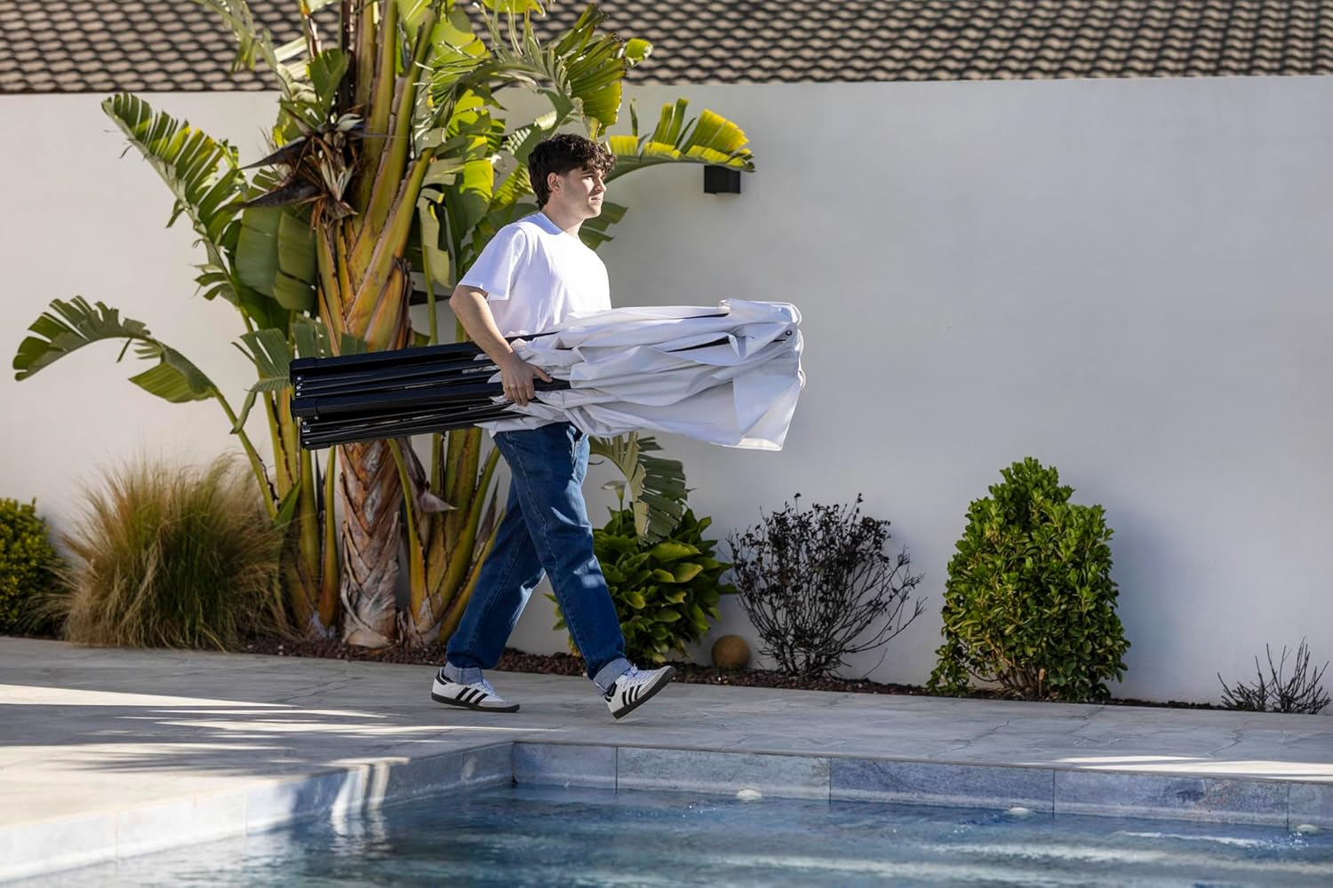 Un hombre con jeans y camiseta blanca lleva una sombrilla plegada junto a una piscina.